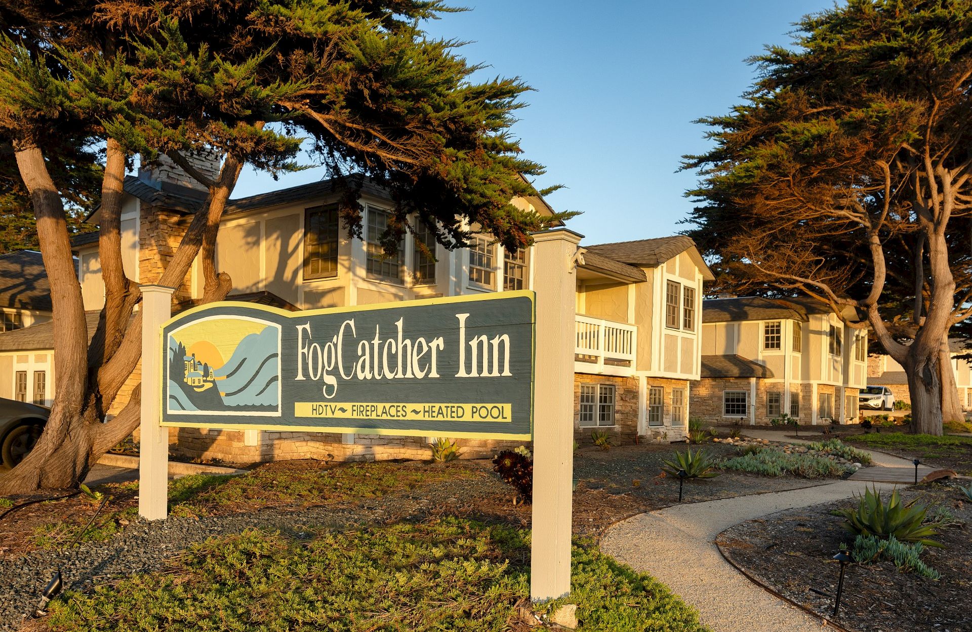 A sign for FogCatcher Inn in front of a seaside-style hotel complex, with a curved path and trees along the walkway.