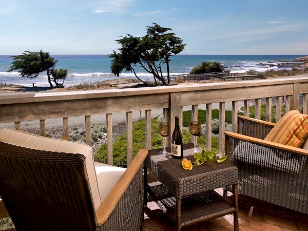 A cozy balcony with wicker chairs, a small table, a bottle of wine, and orange cushions overlooking a rocky coast and blue sea under a clear sky.