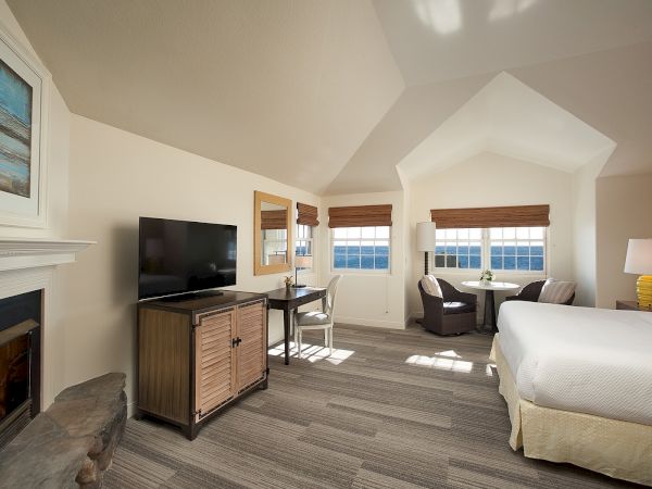 A bright hotel suite with a fireplace, mounted TV, a desk, and a seating area by windows overlooking scenery, plus a bed and nightstand.