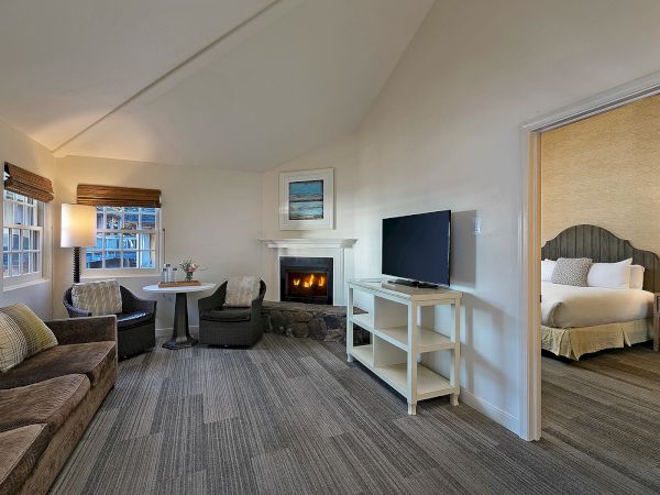 A cozy hotel living area with a fireplace, sofa, two chairs, a small table, and a TV on a stand; a doorway leads to a bedroom with a bed and lamps, in soft neutral tones.