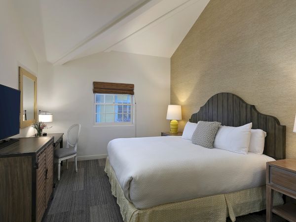 A tidy hotel room with a king bed, two nightstands, a desk and chair, a mounted TV, and soft lighting in neutral tones.