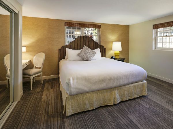 A cozy hotel room with a large bed, nightstands, lamps, a seating area, and a mirror closet, plus windows and wood-toned decor.
