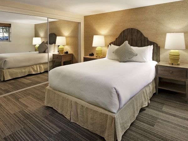 A hotel room with a large bed, mirrored closet wall, two nightstands, lamps, and neutral-toned decor, creating a cozy, symmetrical suite ambiance.