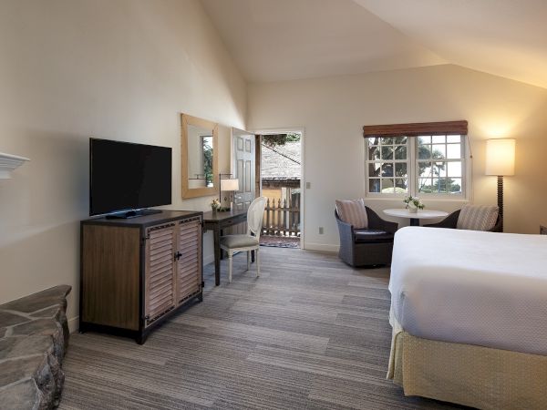 A cozy hotel suite: two beds, a sitting area with sofa, TV on a cabinet, desk, a fireplace, and a balcony view with warm lighting.