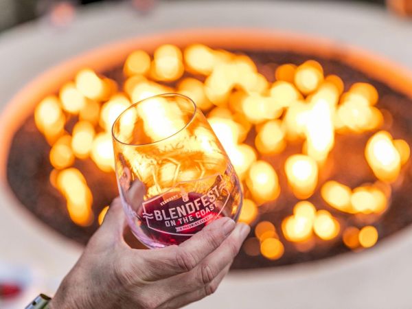 A hand holding a glass with ice and whiskey in front of a glowing fire pit, labeled &ldquo;Blendworks&rdquo; on the glass. ending the sentence.