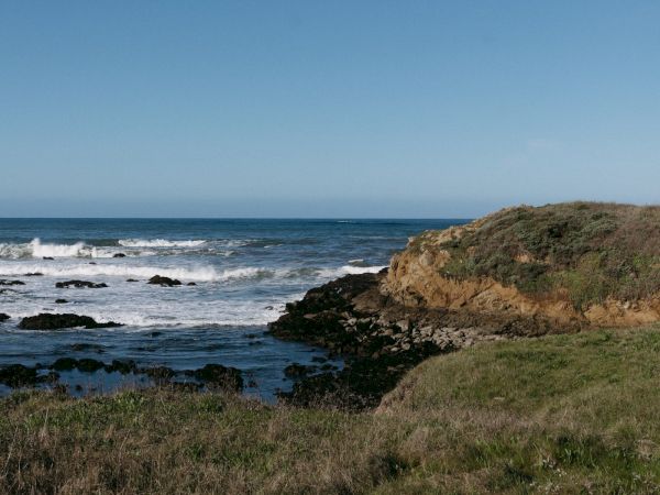 Coastline with rocky shore, grassy foreground, and blue ocean under a clear sky, waves breaking against rocks near a small headland.