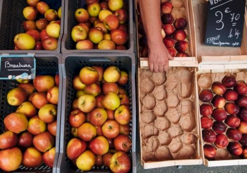 A street market with crates of apples and pears, plus egg cartons and a handwritten price sign on the chalkboard, all arranged in crates.