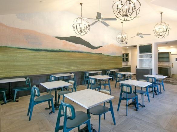 A bright cafe or dining area with turquoise chairs, white tabletops, a mural of hills on the back wall, and modern spherical pendant lights.