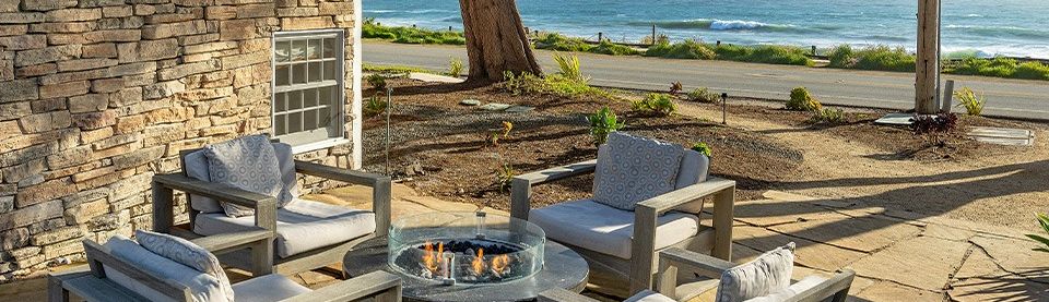 A cozy outdoor seating area with four cushioned chairs around a fire pit on a stone patio, overlooking the ocean.