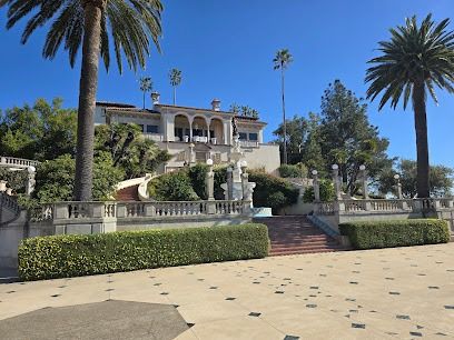 A grand, Mediterranean-style villa atop a landscaped hill with stairs, palm trees, hedges, and a tiled courtyard in sunny weather.