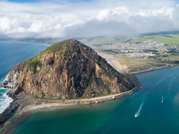 A rocky, cone-shaped island sits in a calm bay, with a breakwater at its base and a town along the coastline beyond, under bright skies.