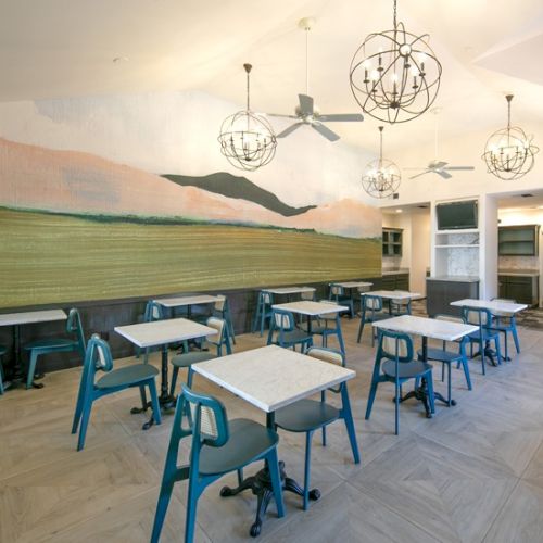 An outdoor classroom setup with empty desks and chairs arranged in rows on a dusty surface, under a clear sky and a mural on a wall.