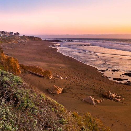 A coastal path on a hilltop during sunset, with sea on one side and rugged dunes, warm orange sky, and soft waves in the distance.