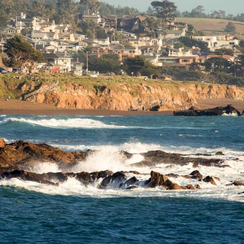 A rocky coastline with waves crashing on a small harbor, blue water, and distant hills under a clear sky.