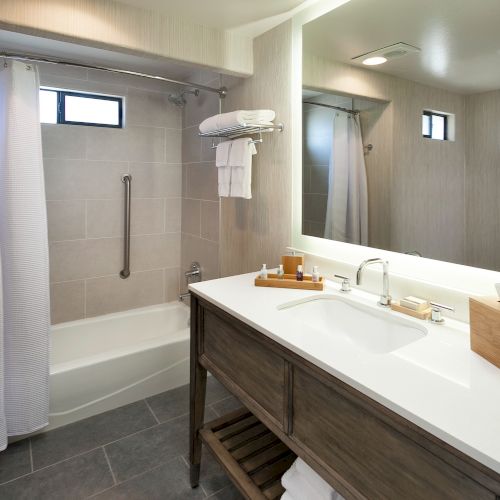 A modern bathroom with a long vanity, twin sinks, large mirror, soft lighting, and a doorway leading to a shower area.