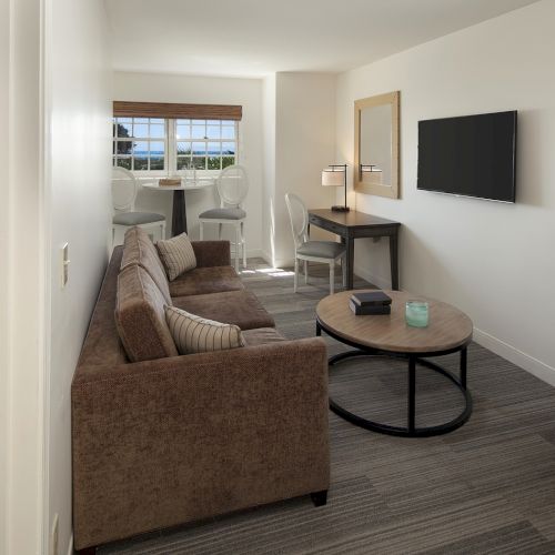 A cozy living room with a couch, coffee table, and wall art; a hallway leads to a bedroom, while warm lighting and a flat-screen decorate the space.