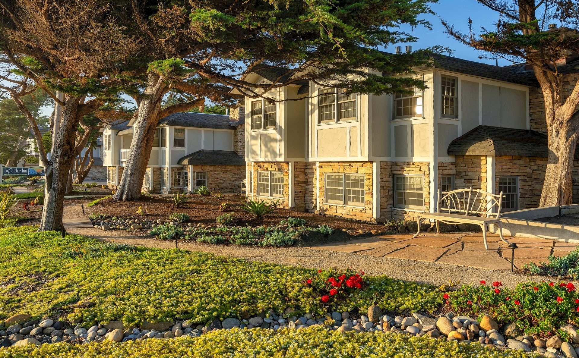 A row of two-story townhomes with stone bases, beige siding, and shutters, set along a neatly landscaped front yard with trees and a walkway.