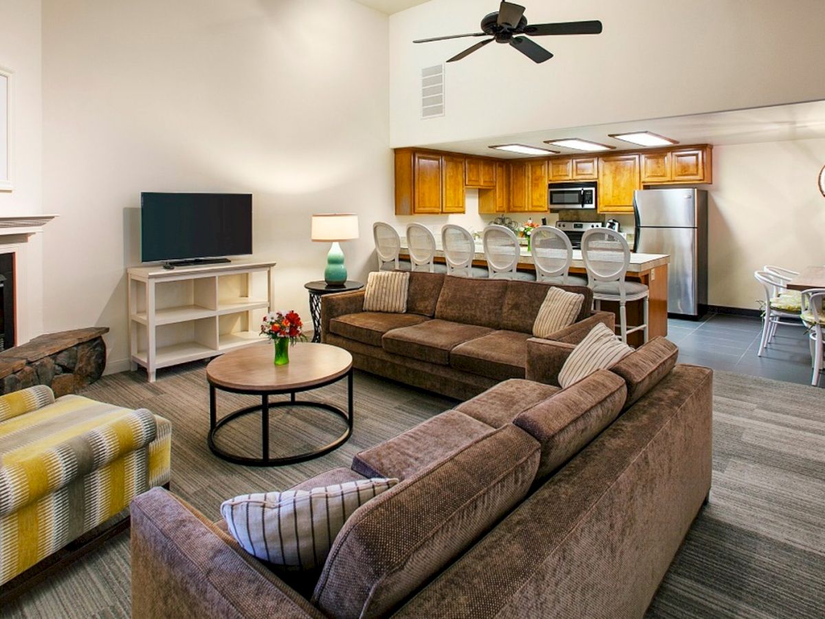 A cozy open-concept living room with a fireplace, brown sectional sofa, yellow plaid chair, coffee table, TV stand, and a bright kitchen with a breakfast bar in the background.