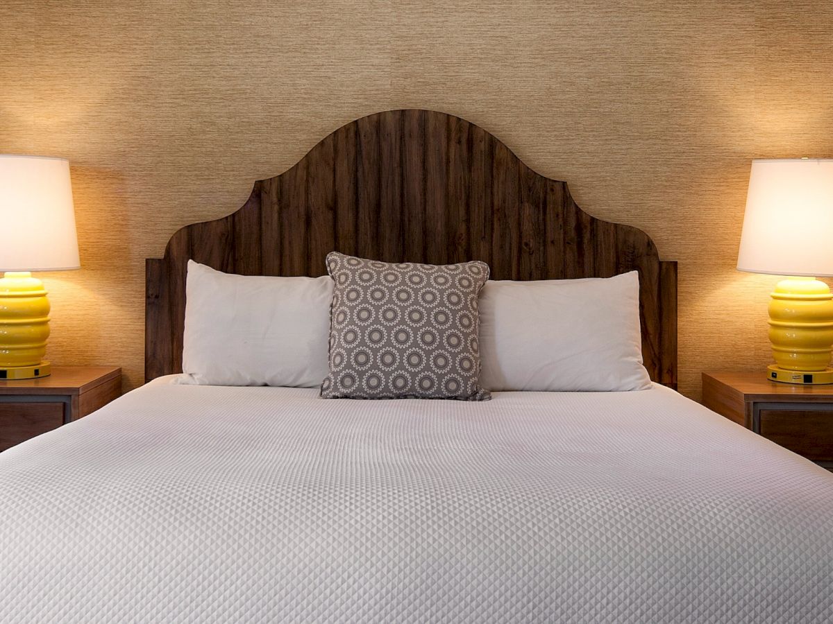 A neatly made bed with a wooden headboard, two bedside tables, matching yellow lamps, and a decorative throw pillow, in a cozy hotel room.