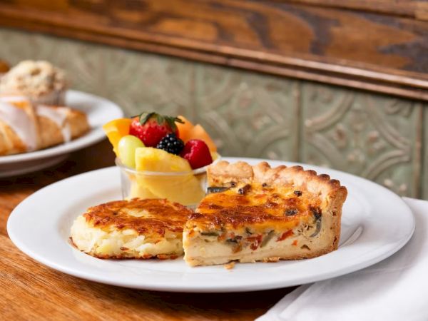 A plate with a slice of quiche or savory pie, another piece, and a fruit cup with berries and mango, on a wooden table.