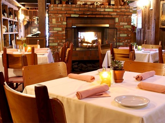 Cozy restaurant with white-tablecloths, lantern light, wooden chairs, and a lit brick fireplace.