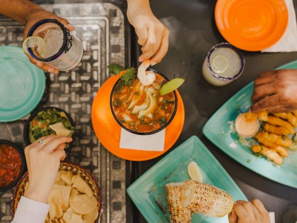 A group of people sharing a colorful meal: tacos, fried foods, and drinks around a table with bright teal plates and an orange dish in the center.