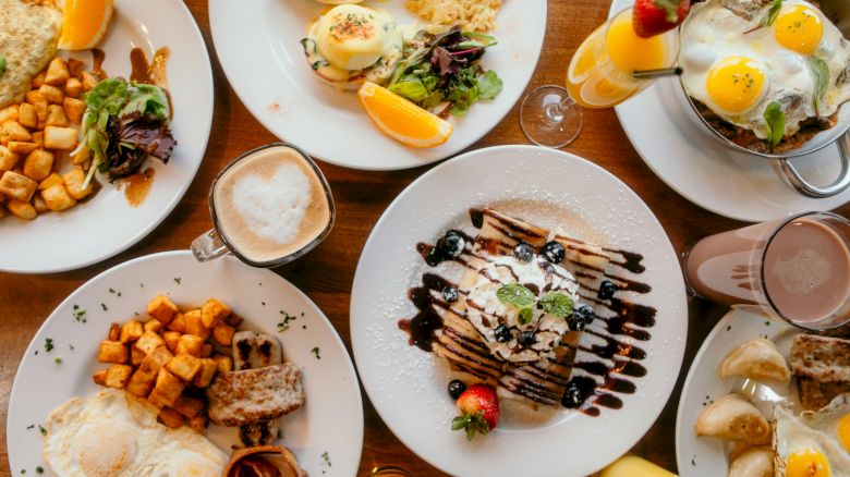 A delicious brunch spread with eggs, toast, potatoes, fruit, pancakes, desserts, and drinks all on a wooden table.