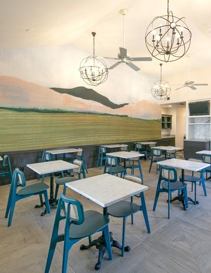 A bright, airy cafe classroom with blue chairs, square tables, a mural of hills and sky, and modern hanging light fixtures.