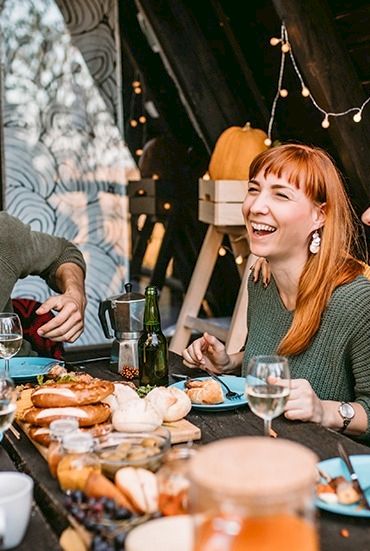 A group of friends sharing a warm meal at an outdoor table, laughing and chatting while coffee and buns are served at sunset.