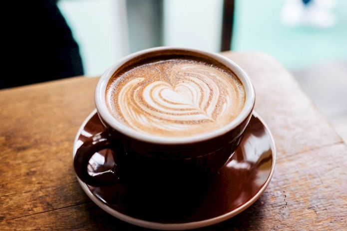 A brown ceramic cup of latte on a wooden table, topped with a heart-shaped foam latte art design, with a saucer underneath.